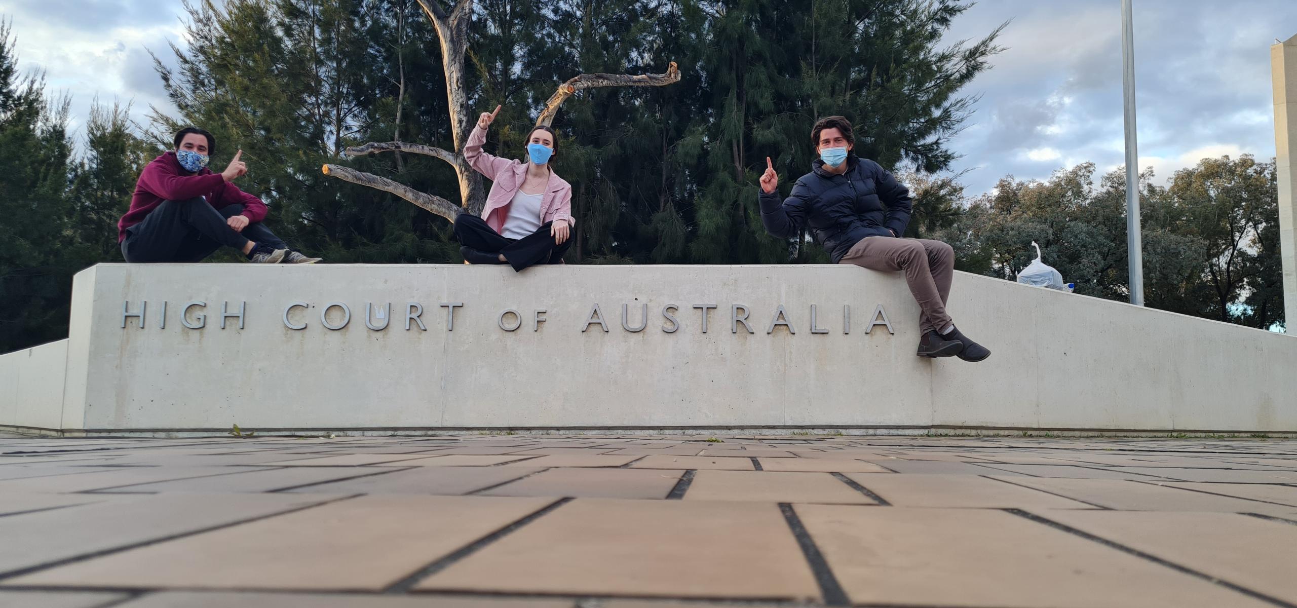 The winning ANU team members Tom Dunbabin, Madeleine McGregor, and Benjamin Durkin, sitting on top of the High Court of Australia sign.